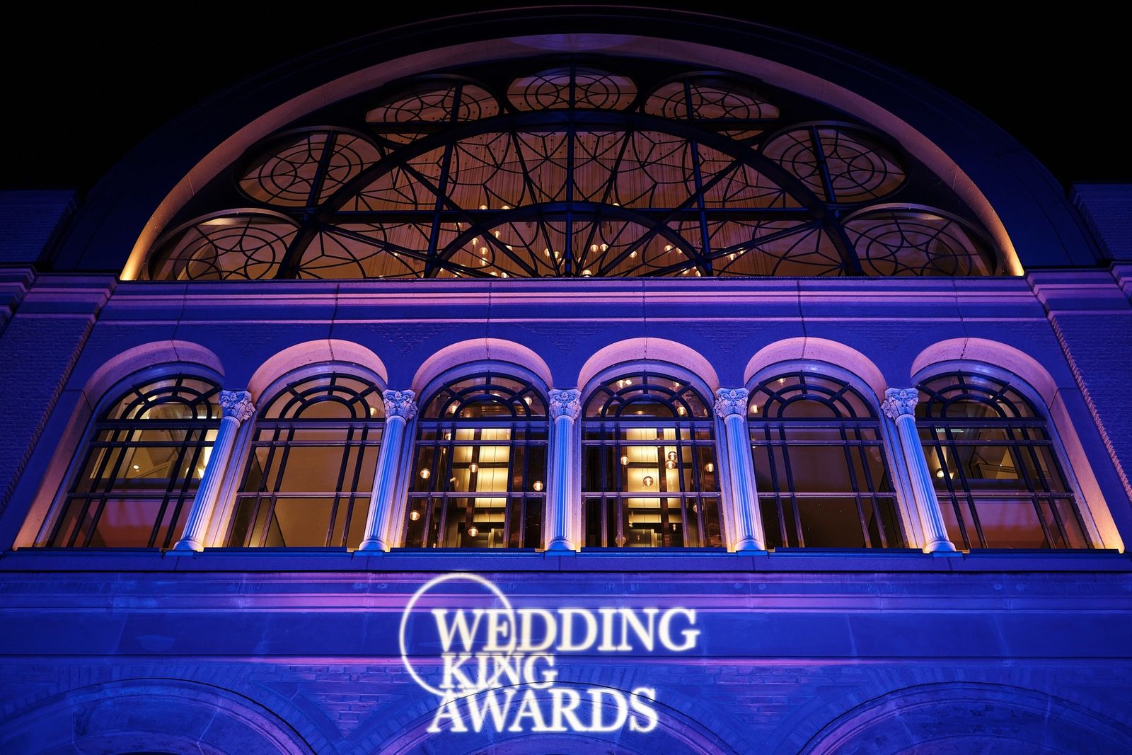 Die Flora Köln bei Nacht, violett angestrahlt, mit projiziertem Wedding-King-Awards-Logo auf der Fassade
